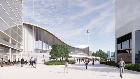 En människoskara vistas på platsen som kantas av en stadion och höghus. På platsen finns en caféterrass och grönområden med träd. Stadion som kantar platsen har ett välvt tak med breda trappor som leder till ingångsnivån.