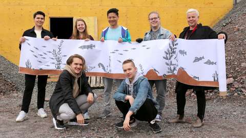 Kuvassa opiskelijat seisovat tunnelin edessä ja pitävät ylhäällä kuvaa heidän tekemästään seinätaiteesta.