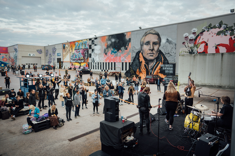 Ihmisiä ulkoilmalavan edustalla. Lavalla muusikkoja. Taustalla suurikokoisia seinämuraaleja.