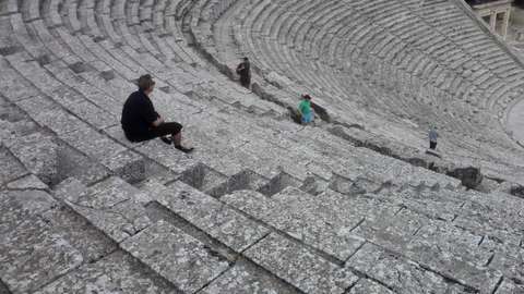 Sami kuvattuna takaapäin, alaspäin syvenevillä portailla istuen. 