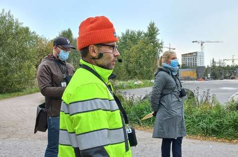 Kolme tapahtuman vetäjää puhumassa mikrofoneihin ulkona. Taustalla puita, Finnoon uusia kerrostaloja ja nostureita.