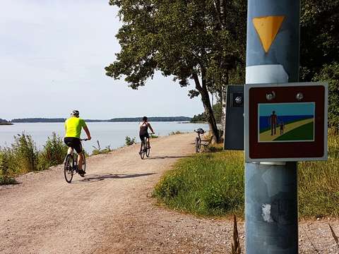 Kuvassa näkyy pyöräilijöitä rantaraitilla sekä rantaraitin logo Westendissä.