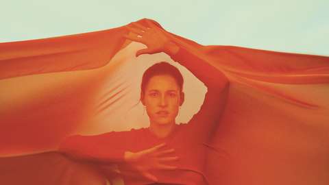 Punaiseen, suureen huiviin kietoutunut nainen pitää käsiään kohotettuina.