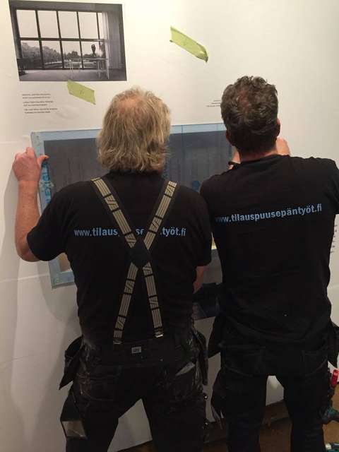 Kaksi miestä selin kameraan asentamassa pleksiä seinään.