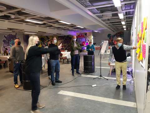About ten persons are standing and discussing with facemasks on their face in front of a wall with post it notes.