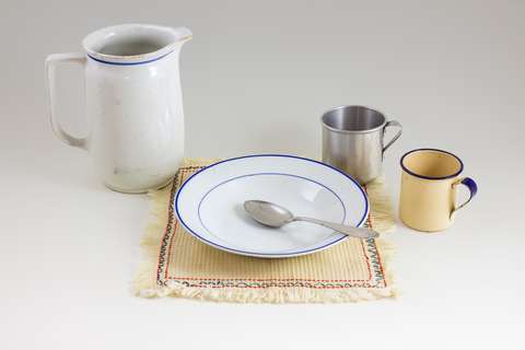 Ruokailuvälineitä pöydällä. Kannu, lautanen liinan päällä, lusikka ja kaksi mukia.