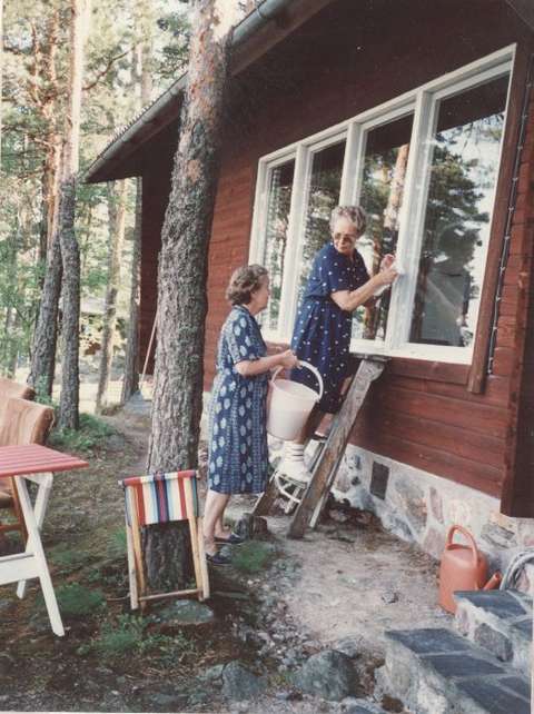 Kaksi naista pesee kesämökin ikkunoita kesäisessä kelissä.