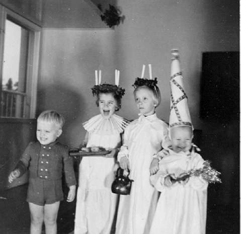 Neljä pientä lasta juhlaan pukeutuneena Lucia-kulkueessa. Mustavalkokuva.