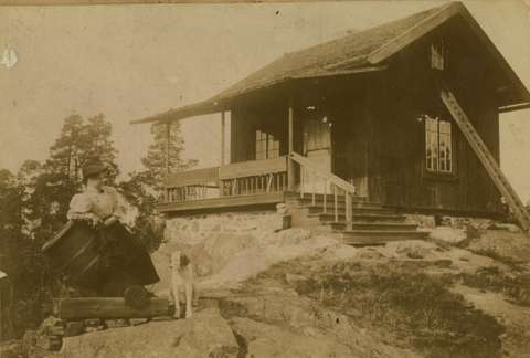 Nainen nojaa mörssäriin, taustalla mökki.