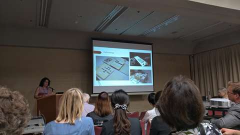 Kuvassa ihmisiä luokkahuoneessa katse luennoitsijaan sekä esitelmän kuvaesitykseen.