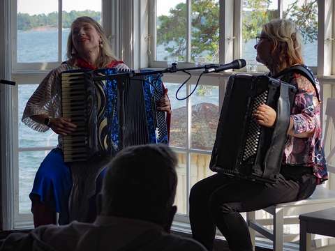 Kaksi naista soittaa harmonikkaa kuistilla, taustalla kesäinen merimaisema.