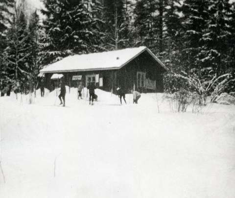 Pieni joukko hiihtäjiä torpan edessä lumisessa metsämaisemassa, mustavalkoinen valokuva.
