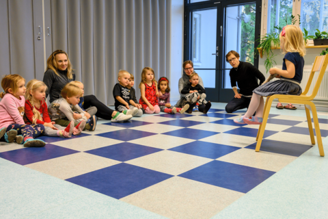 Children and three instructors are sitting on the floor. One child is sitting on a chair in front of others looking at one of the instructors.
