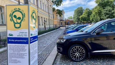 Pysäköintikokeiluja mainostava kyltti ja pysäköityjä autoja kivipäällysteisellä torilla.