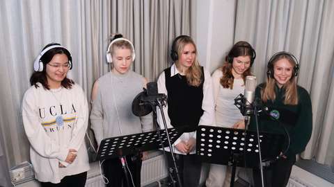 Viisi laulajaa seisoo äänitysmikrofonien takana. 