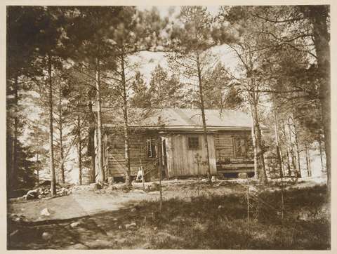 Puusta rakennettu tupa mäen laella metsikön ympäröimänä, mustavalkoinen valokuva.