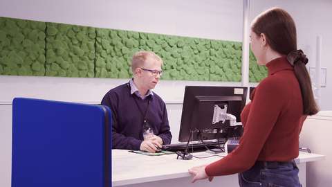 Nainen seisoo palvelutiskin äärellä, ja tiskin takana istuu asiointipisteen työntekijä.