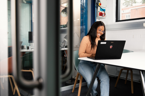 Valokuva, jossa nuori nainen työskentelee tietokoneella. Toimistoympäristö.