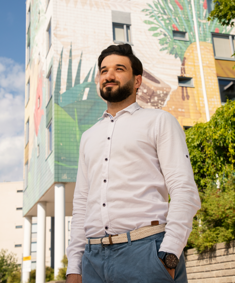 Valokuvassa mies seisoo ja hymyilee. Taustalla kerrostalo, jossa muraali.