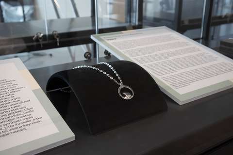 Vitriinissä on mustalla tyynyllä riipus, jonka molemmin puolin on tekstiä sisältävät taulut.