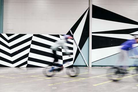 Kaksi vauhdikkaasti pyöräilevää nuorta suuren seinämaalauksen edessä.