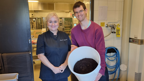 Kaksi henkilöä seisoo keittiössä. Toisella on kädessä 20 litran astia, jossa on käytettyjä kahvinporoja.