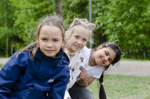 Kolme lasta katsoo kameraan ulkona. Kahdella on kissan korvat päässä.