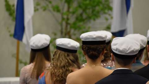 Ylioppilaita istumassa salissa lakki päässä. Opiskelijat on kuvattu selkäpuolelta.