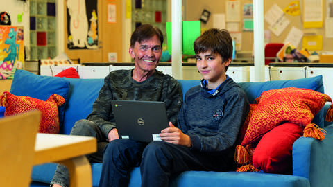  A man and a boy sit on a blue couch in a colorful classroom.