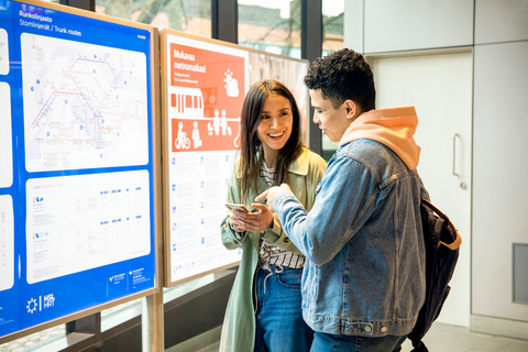 Kaksi ihmistä metroaseman opastintaulun edessä katsomassa kännykkää.