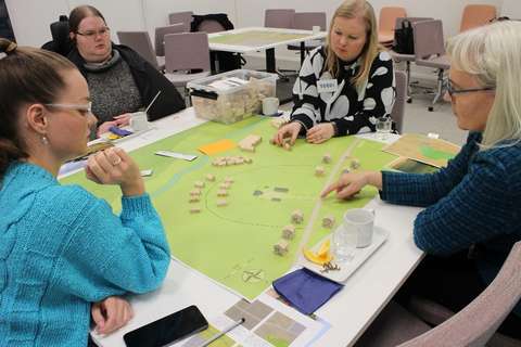 Deltagare i verkstaden undersöker material på bordet. Materialet består av små träklosshus och spelplattform är en karta över en påhittad stadsdel.
