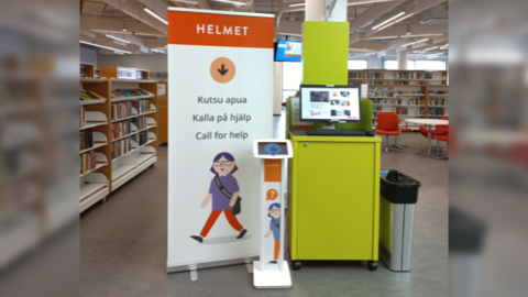 Kuvassa jalustan päällä oleva tabletti sekä banderolli, jossa on Helmet-logo ja lukee Kutsu apua, Kalla på hjälp, Call for help. Banderollissa ja tabletin jalustassa on piirroskuva kirjastotyöntekijästä. 