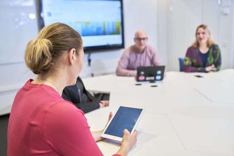 Neljä henkilöä keskustelee ja katsoo tietokonetta ja tablettia neuvottelupöydän ääressä.