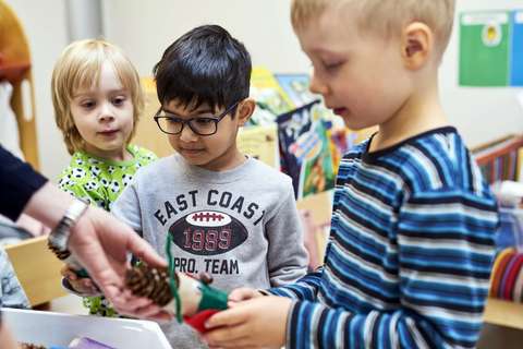 Kolme Tuomarilan päiväkodin lasta tutkivat yhdessä opettajan kädessä olevaa askartelua.