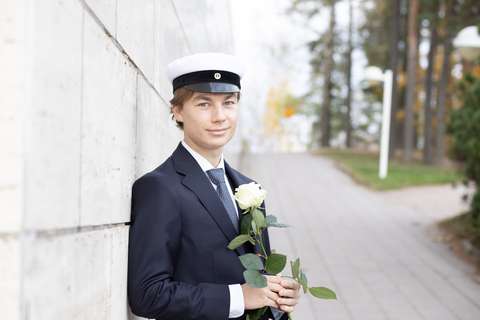 Nuori mies ylioppilaslakki päässään. Taustalla puistoa ja talon tiiliseinä.