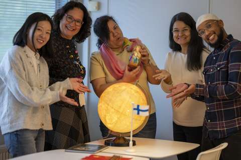 Kulttuuriohjaajat poseeraavat kameralle. Keskimmäinen henkilö pitää kädessään esinettä. Pöydällä on karttapallo ja Suomen lippu.