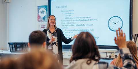 Opettaja seisoo luokan edessä screeni taustallaan. Opiskelijat viittaavat.