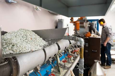 Grå plast i småbitar i matningstråget på en stor industriell maskin, i bakgrunden en man med hörselskydd.