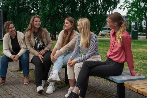 Viisi nuorta istuu kesällä penkillä puistossa ja katsovat naureskellen toisiaan.