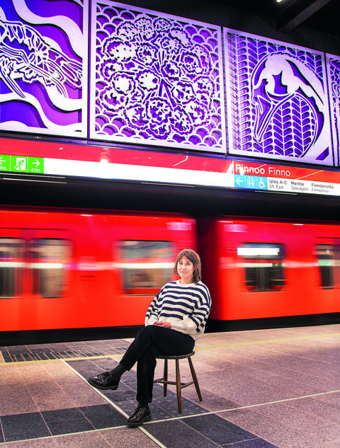 Leena Nio istuu tuolilla Finnoon metroaseman metrolaiturilla ja katsoo hymyillen kameraan. Metrojuna on liikkeellä Nion takana. Kuvan yläosassa näkyy Nion taideteos Haavekuvia.