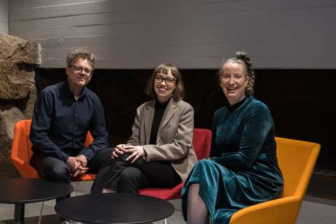 Kolme hymyilevää henkilöä istuu nojatuoleissa pöydän ääressä.