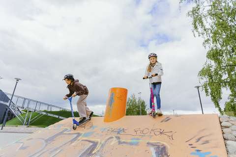 Valokuva. Kaksi lasta skuuttaa. Vasemman puoleinen lapsi on laskemassa kummulta alas ja oikeapuoleinen lapsi on kummun päällä.