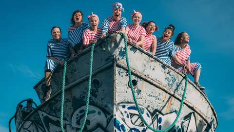 Kahdeksan raidallisiin asuihin pukeutunutta henkilöä seisoo ruostuneen veneen etuosassa. 