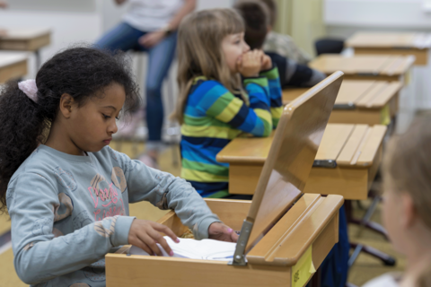 Lapset istuvat luokkahuoneessa pulpeteissaan. Yksi lapsi tutkii pulpettinsa sisältöä.