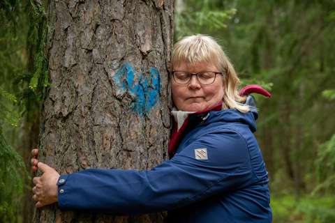 Nainen halaa suurikokoista puuta silmät kiinni, puun runkoon on maalattu sininen sydän.