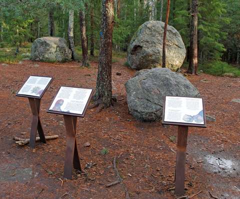 Luontopolkuopasteita kuluneessa maastossa, taustalla suuria siirtolohkareita.