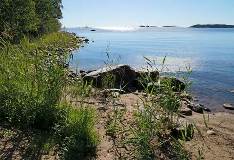 Rantamaisema hiekkarannasta, kivikosta ja rantakasvillisuudesta, taustalla sininen meri ja horisontissa saaria