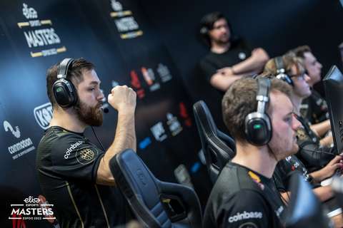 Kuvassa näkyy miehiä istumassa rivissä tietokoneruutujen edessä. Kaikilla kuulokkeet päässään. Takana näkyy mies, joka tuulettaa kädellään.