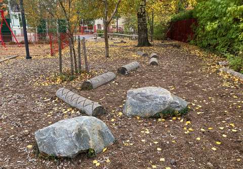 Päiväkodin piha, jolla on suurikokoisia kiviä, puunrunkoja ja kasvillisuutta.