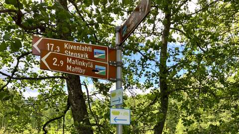 Kuvassa näkyy metallinen tolppa, jossa on ruskeita tienviittoja. Niiden alapuolella on kaksi valkopohjaista kylttiä, joissa on vihreä laiva ja pyörä sekä nuoli oikealle tai vasemmalle. Taustalla näkyy vehreitä puita.